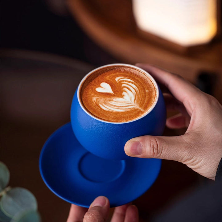 Overhead close-up of woman holding Klein blue ceramic cups with latte art, elegant stylish coffee setup