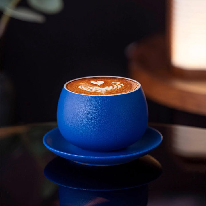 Klein blue ceramic coffee cup with latte art on table, elegant refined setup with ambient lighting