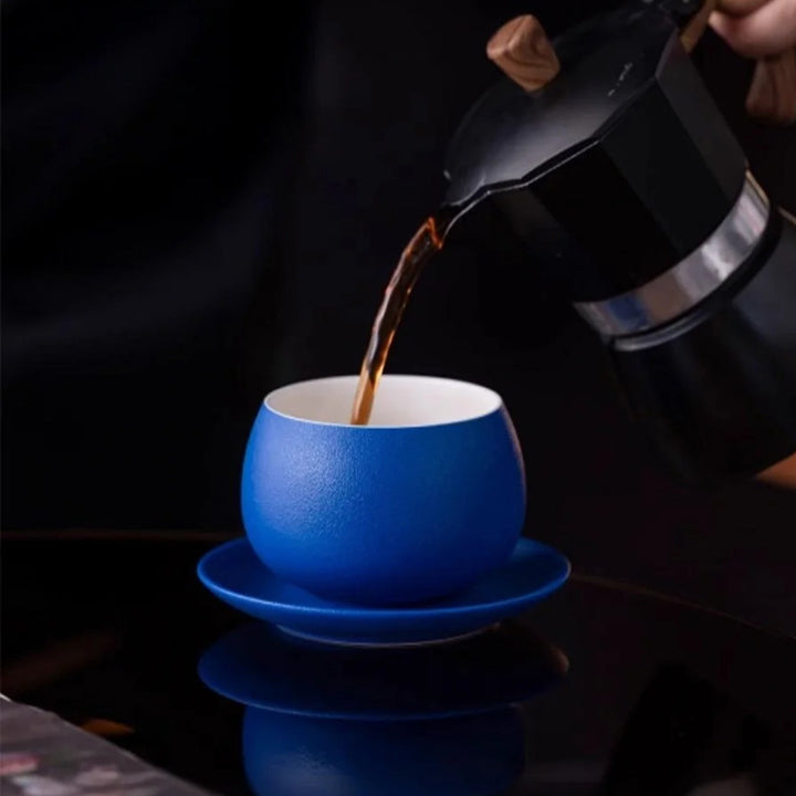 Klein blue ceramic coffee cup being filled from coffee pot on dark background, elegant classic coffee moment